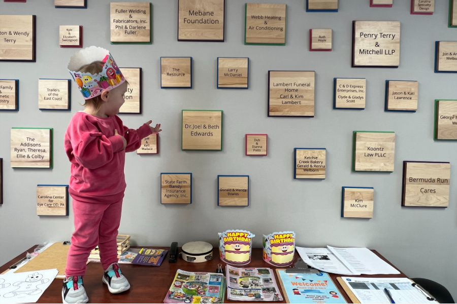 A pre-schoooler wearing her birthday crown stands next to the Smart Start Wall of honor. 